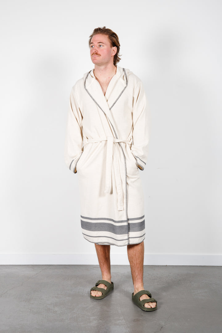 A man standing wearing a medium length beige hooded belted terry robe with gray stripes, and pockets. 