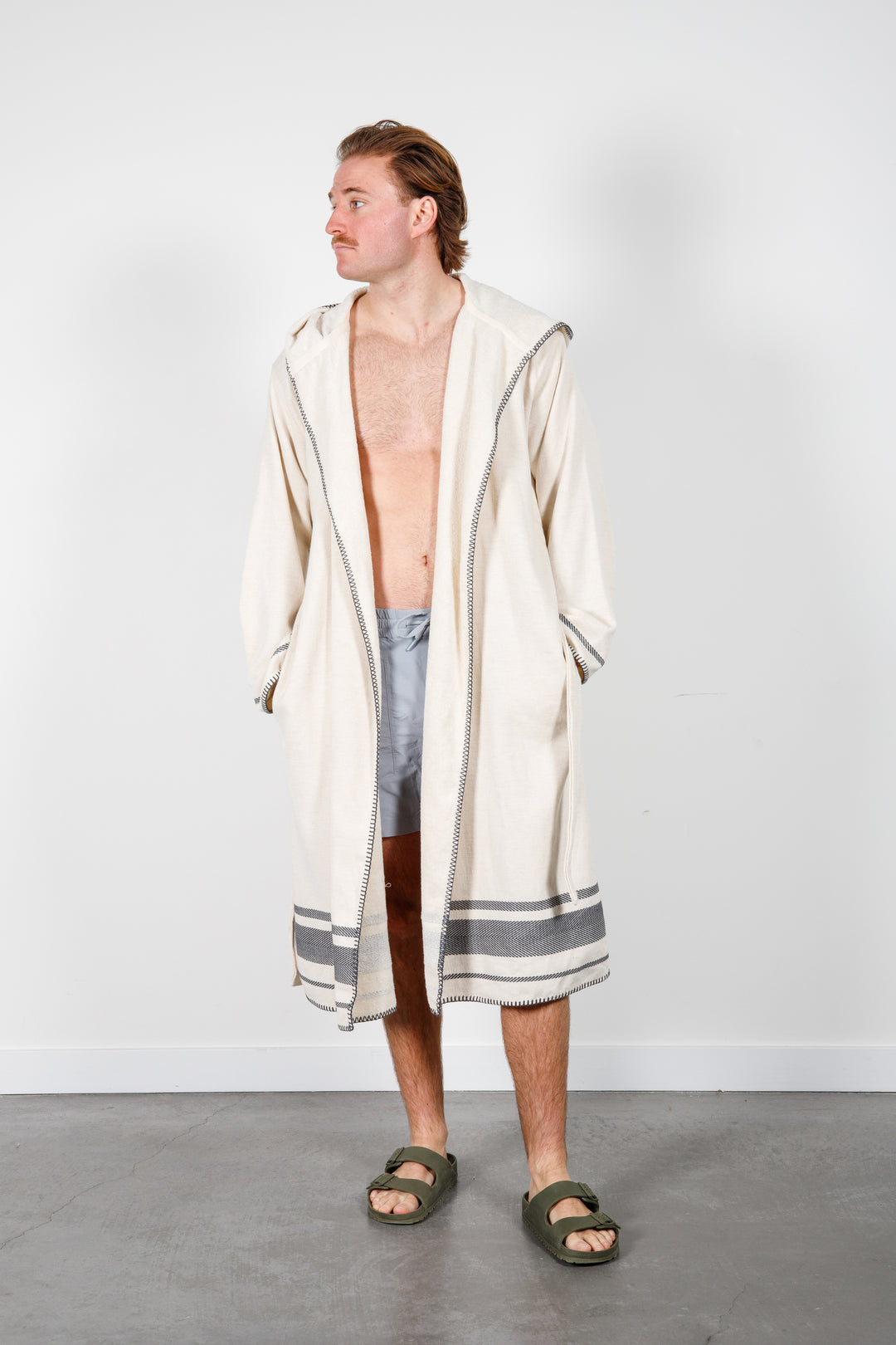 A man standing wearing a medium length beige hooded belted terry robe with gray stripes, and pockets. 