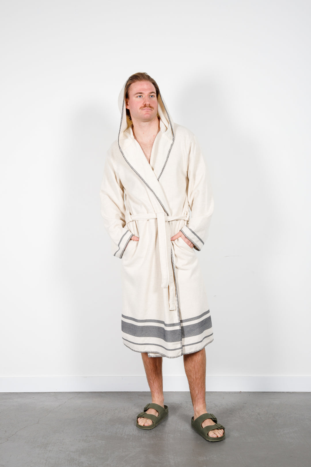 A man standing wearing a medium length beige hooded belted terry robe with gray stripes, and pockets. Hood up