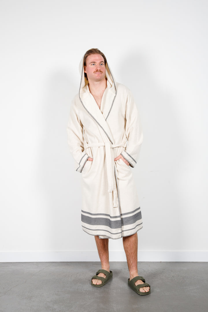 A man standing wearing a medium length beige hooded belted terry robe with gray stripes, and pockets. Hood up