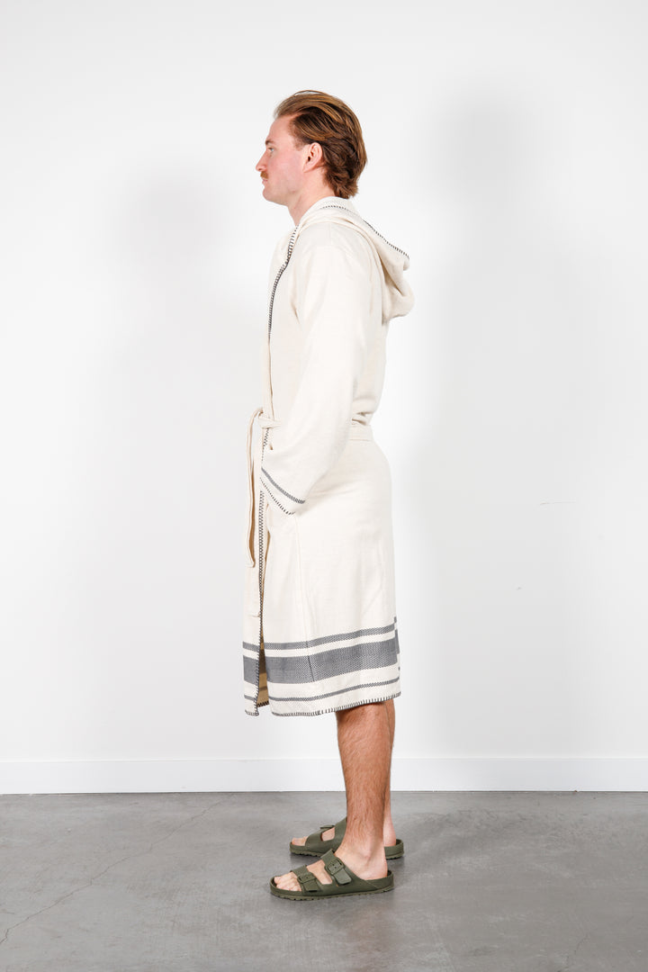 A man standing wearing a medium length beige hooded belted terry robe with gray stripes, and pockets. Hands in pockets