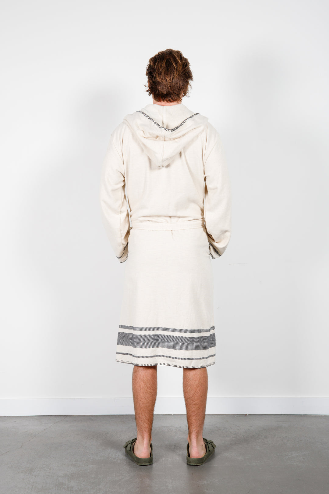 A man standing backwards wearing a medium length beige hooded belted terry robe with gray stripes, and pockets. 