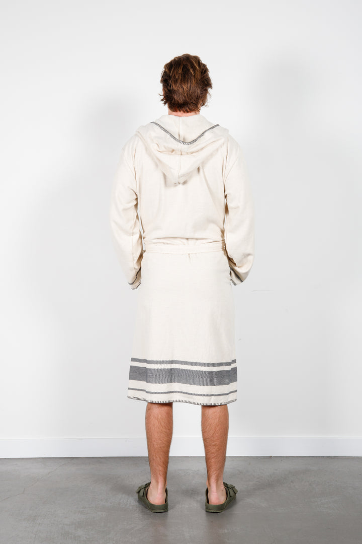 A man standing backwards wearing a medium length beige hooded belted terry robe with gray stripes, and pockets. 