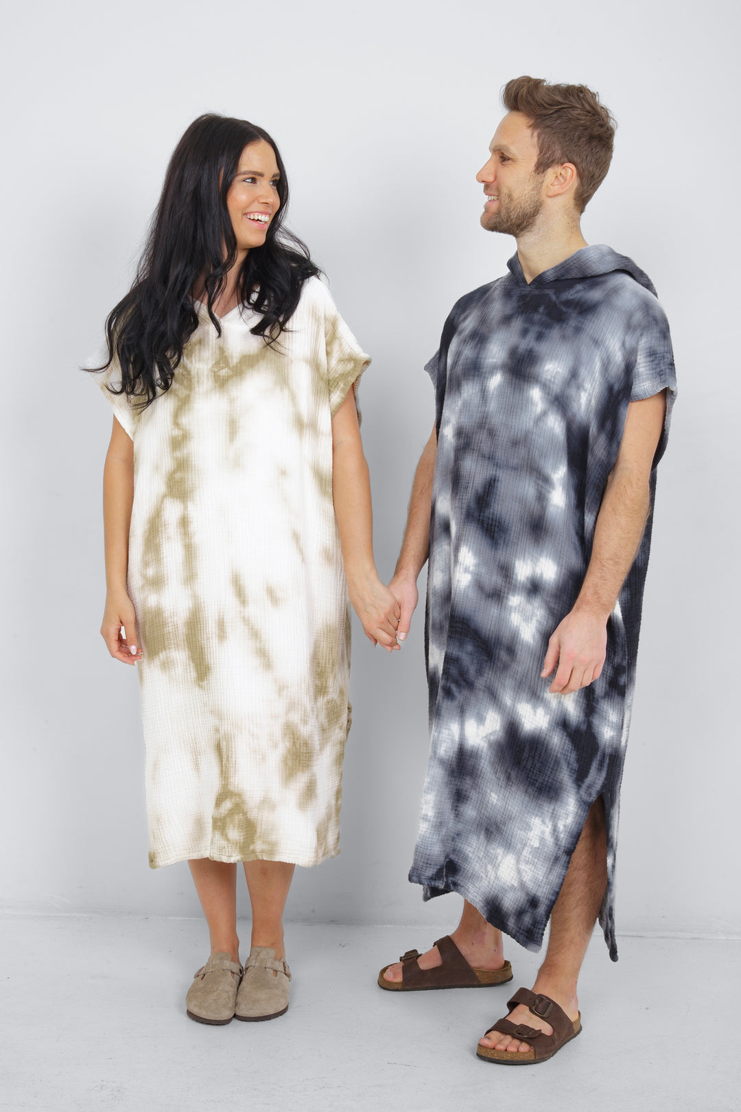 A man and a woman standing side by side wearing hooded, tie-dye muslin surf ponchos with side slits in different colors.  #colour_khaki-tie-dye