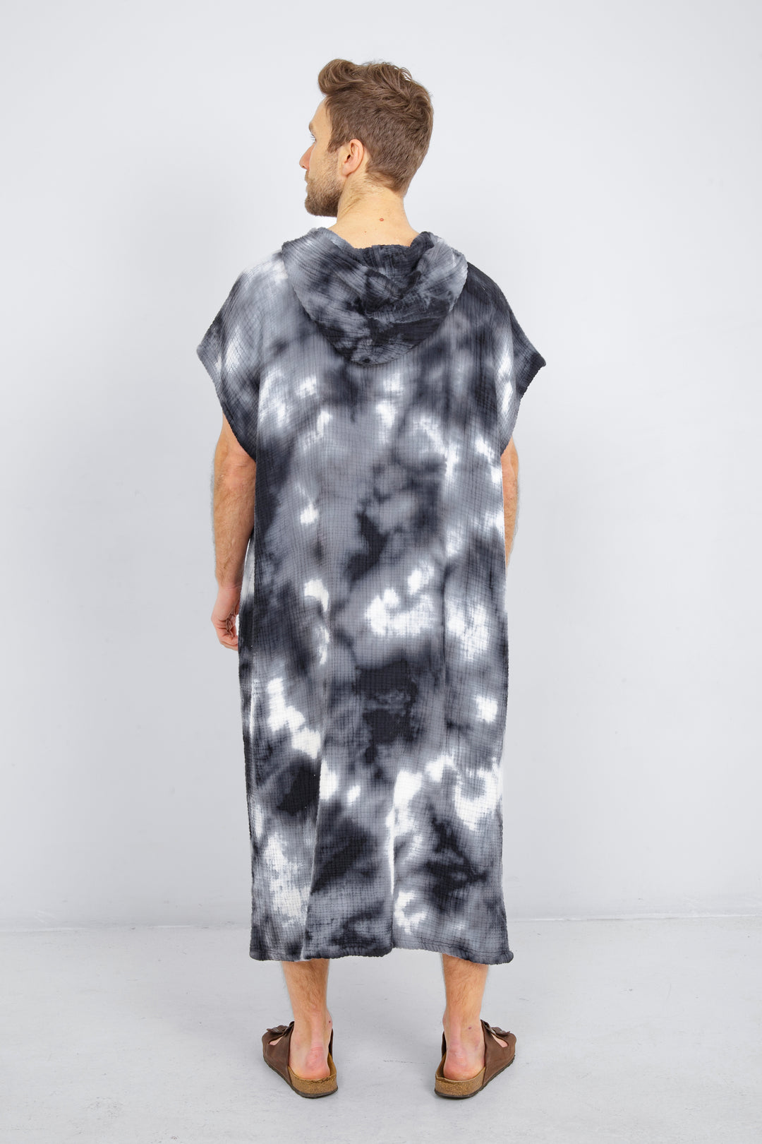 A man standing backwards wearing a dark blue and white hooded, tie-dye muslin surf poncho with side slits  #colour_ocean-tie-dye