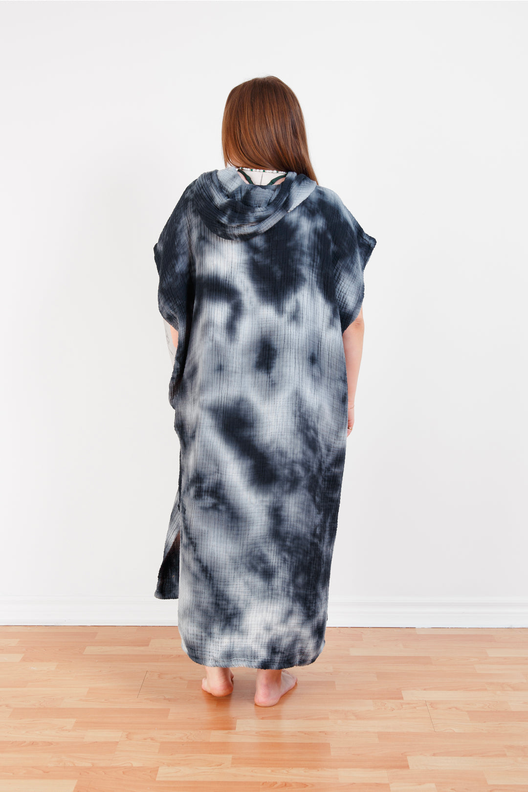 A woman standing backwards wearing a dark blue and white hooded, tie-dye muslin surf poncho with side slits  #colour_ocean-tie-dye