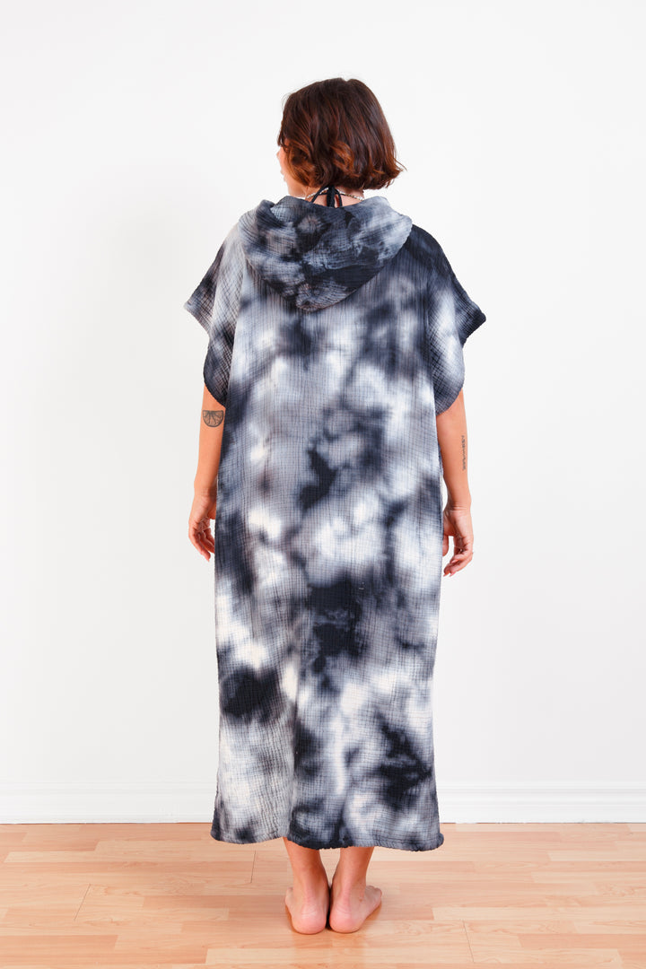 A woman standing backwards wearing a dark blue and white hooded, tie-dye muslin surf poncho with side slits  #colour_ocean-tie-dye