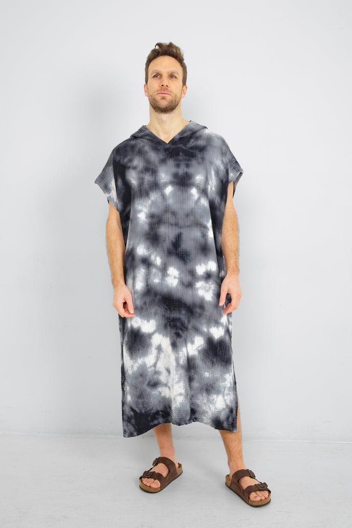 A man standing wearing a dark blue and white hooded, tie-dye muslin surf poncho with side slits  #colour_ocean-tie-dye