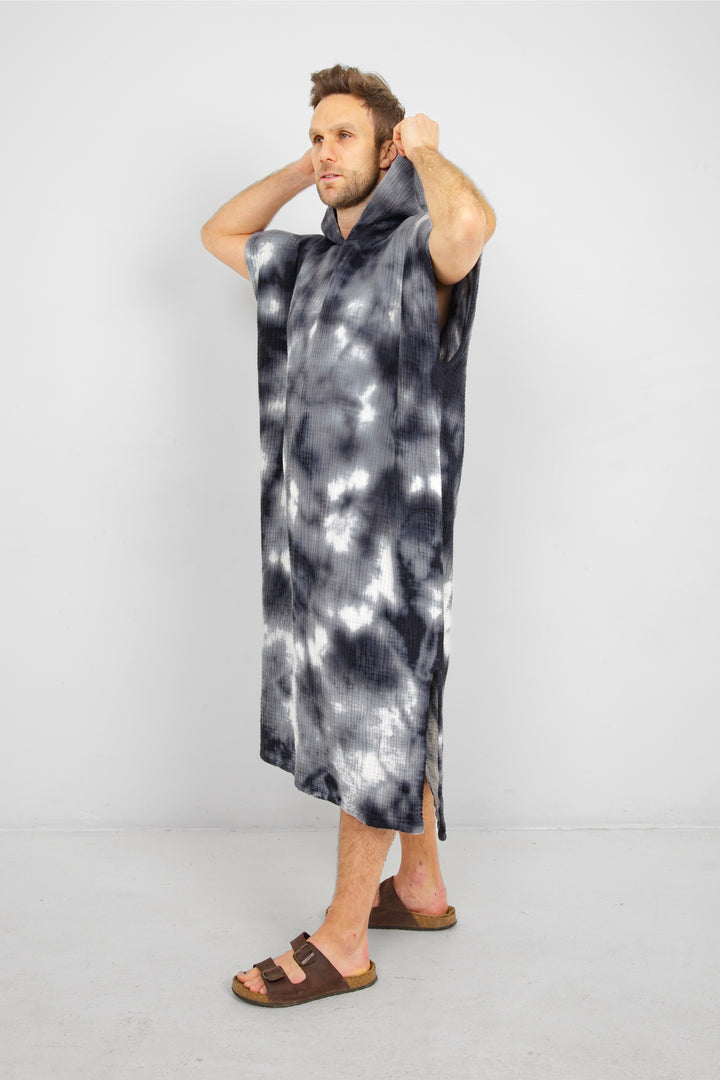 A man standing wearing a dark blue and white hooded, tie-dye muslin surf poncho with side slits  #colour_ocean-tie-dye