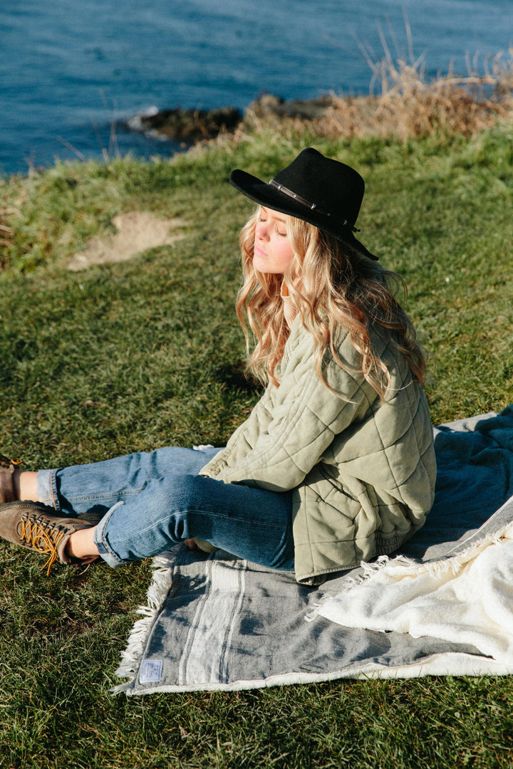 A blond girl in jeans and a light green quilted jacket and a black brimmed hat sitting on a dark-grey fleece-lined Turkish throw with eyelash fringe, and simple stylized white stripes, outdoors in the sun on grass near the ocean #colour_granite
