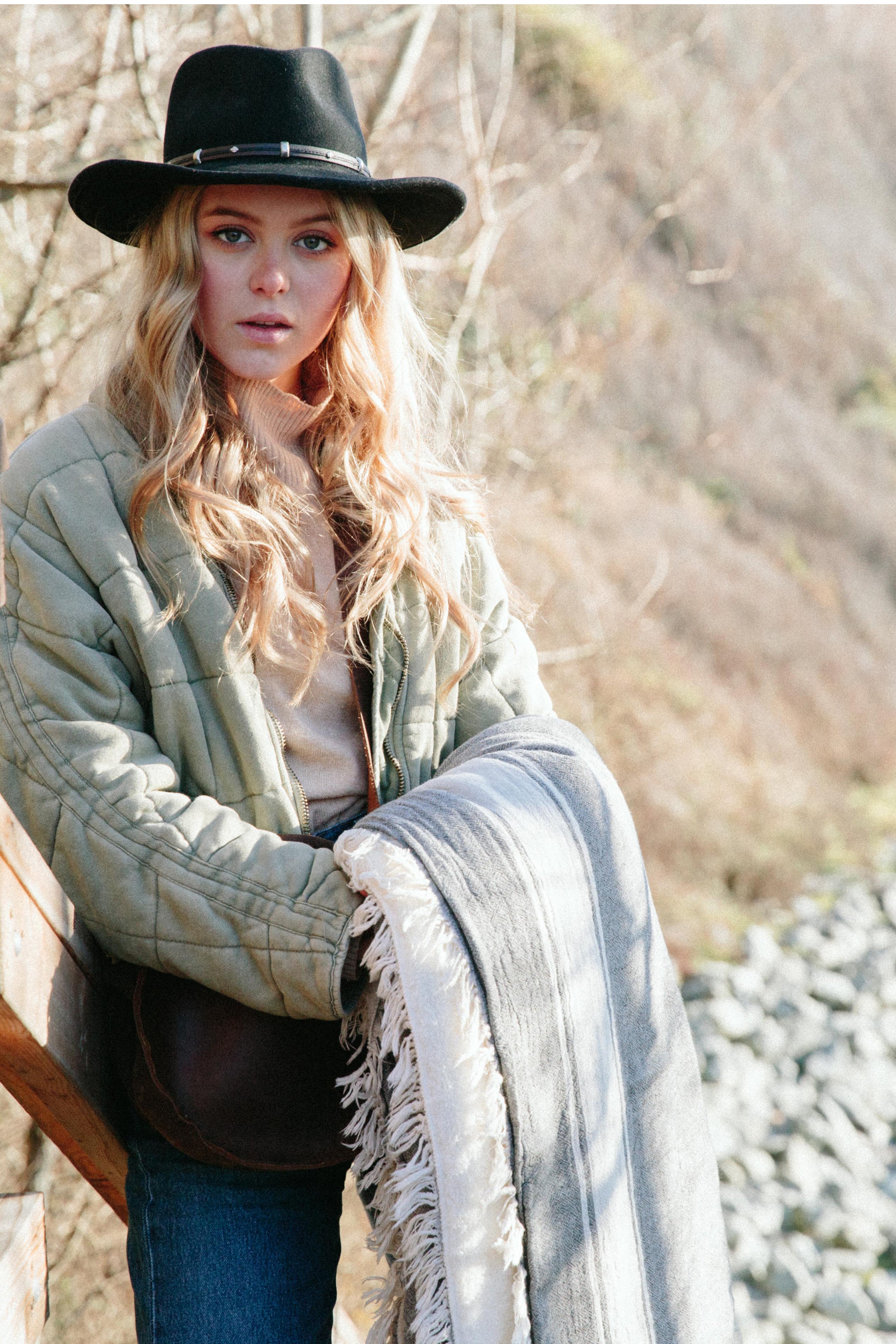 A blond girl in jeans and a light green quilted jacket and a black brimmed hat is holding a dark-grey fleece-lined Turkish throw with eyelash fringe, and simple stylized white stripes, outdoors #colour_granite