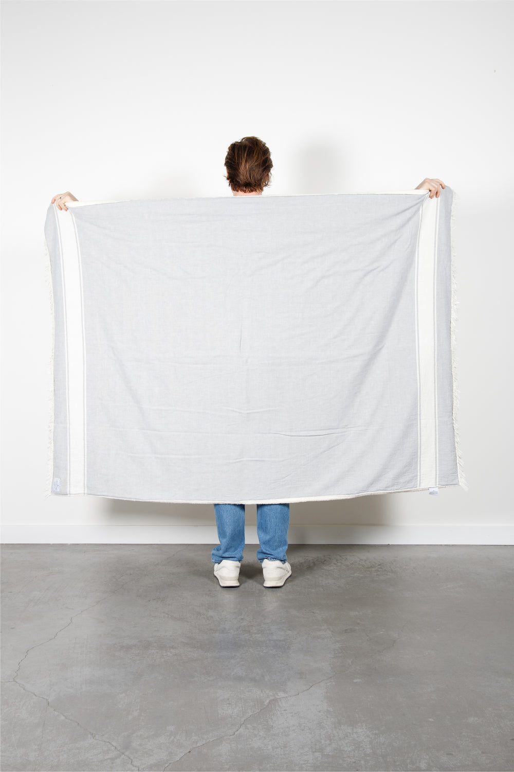 A man wearing jeans is holding upa light-gray fleece-lined Turkish throw with eyelash fringe, and simple, stylized white stripes, in a room with a white wall and concrete floor. #colour_pewter