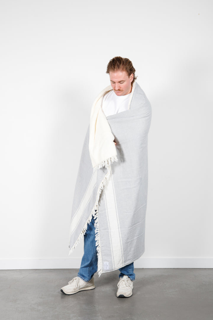 A man wearing jeans and a white t-shirt is wrapped in a light-gray fleece-lined Turkish throw with eyelash fringe, and simple, stylized white stripes, in a room with a white wall and concrete floor. #colour_pewter