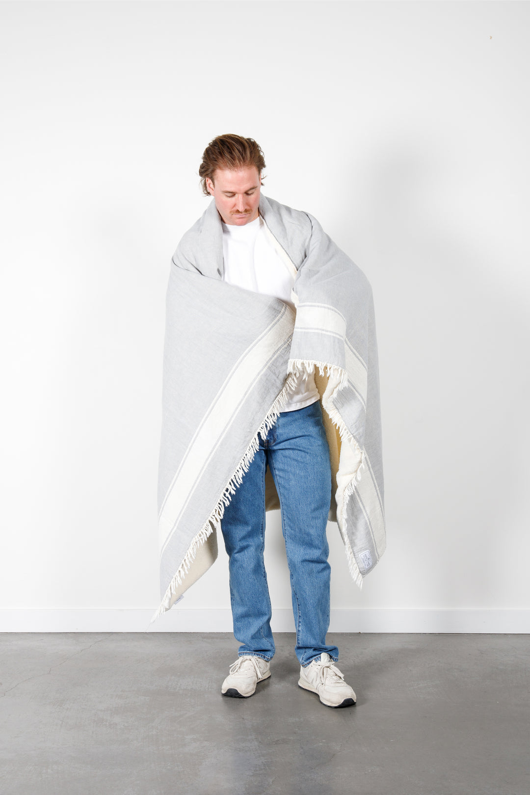 A man wearing jeans and a white t-shirt is wrapped in a light-gray fleece-lined Turkish throw with eyelash fringe, and simple, stylized white stripes, in a room with a white wall and concrete floor. #colour_pewter