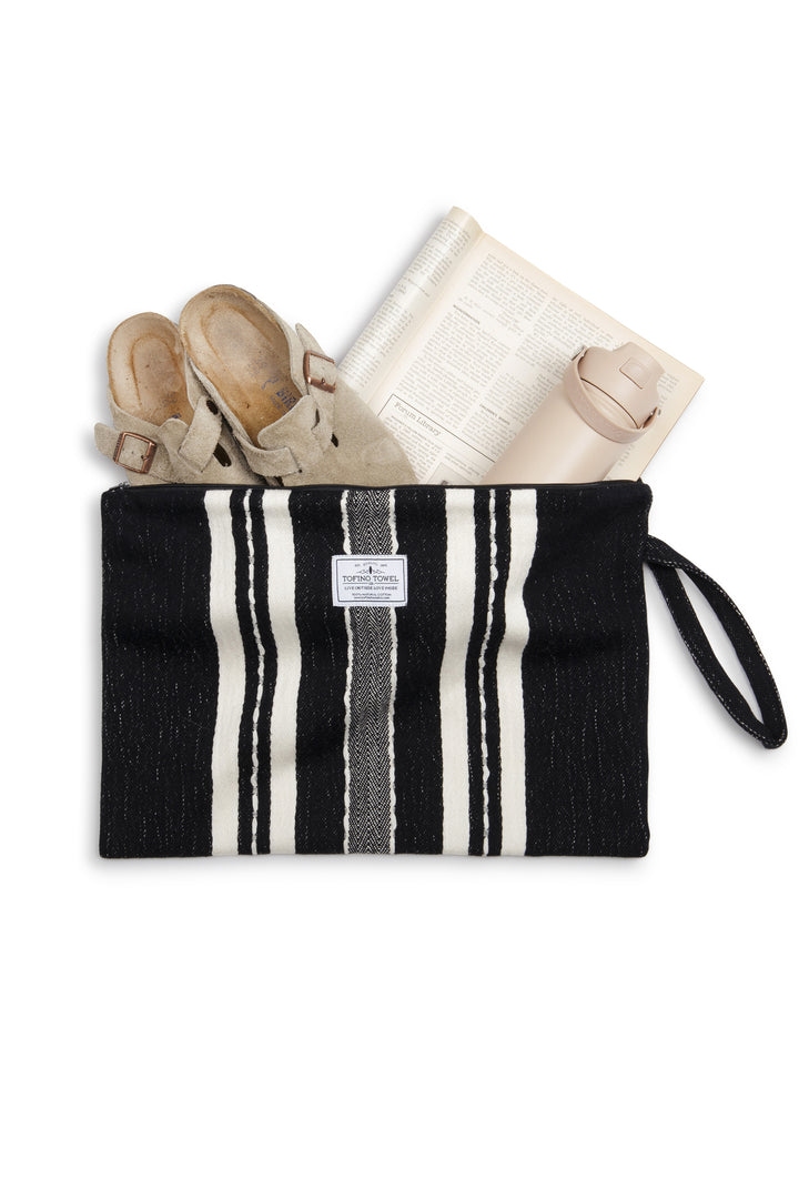 Large Black zip up pouch with white stripes and a wrist handle containing a pair of shows a water bottle and a magazine on a white background