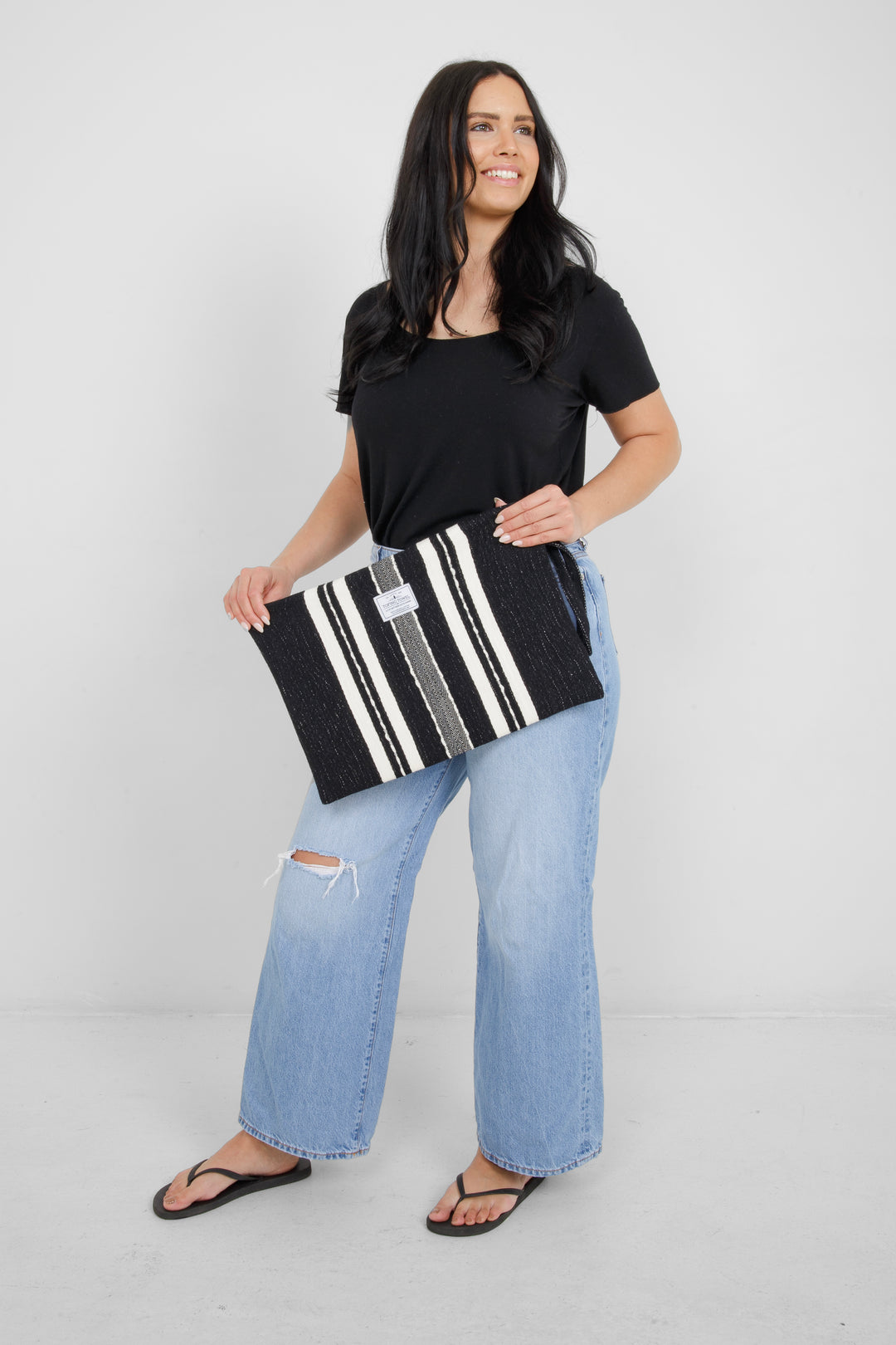 Woman holding in light blue jeans and a black t shirt holding a Large black zip up pouch with white stripes and a wrist handle on a white background