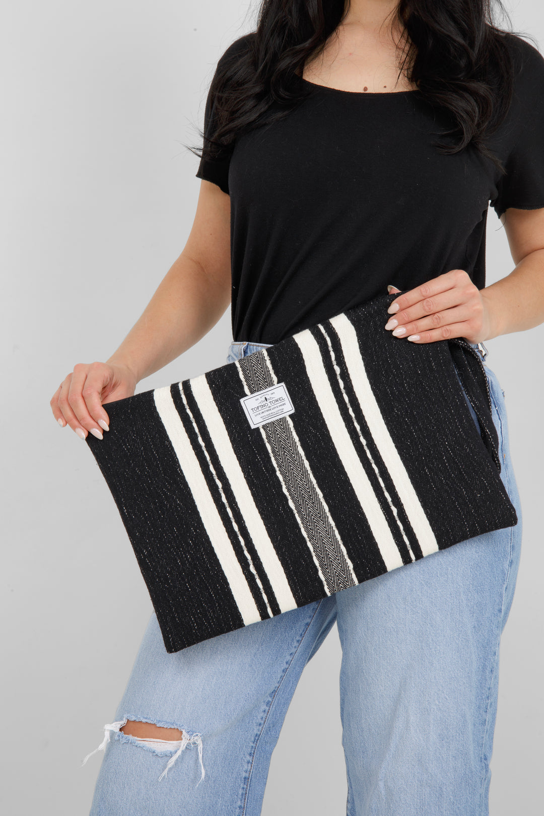 Woman holding in light blue jeans and a black t shirt holding a Large black zip up pouch with white stripes and a wrist handle on a white background
