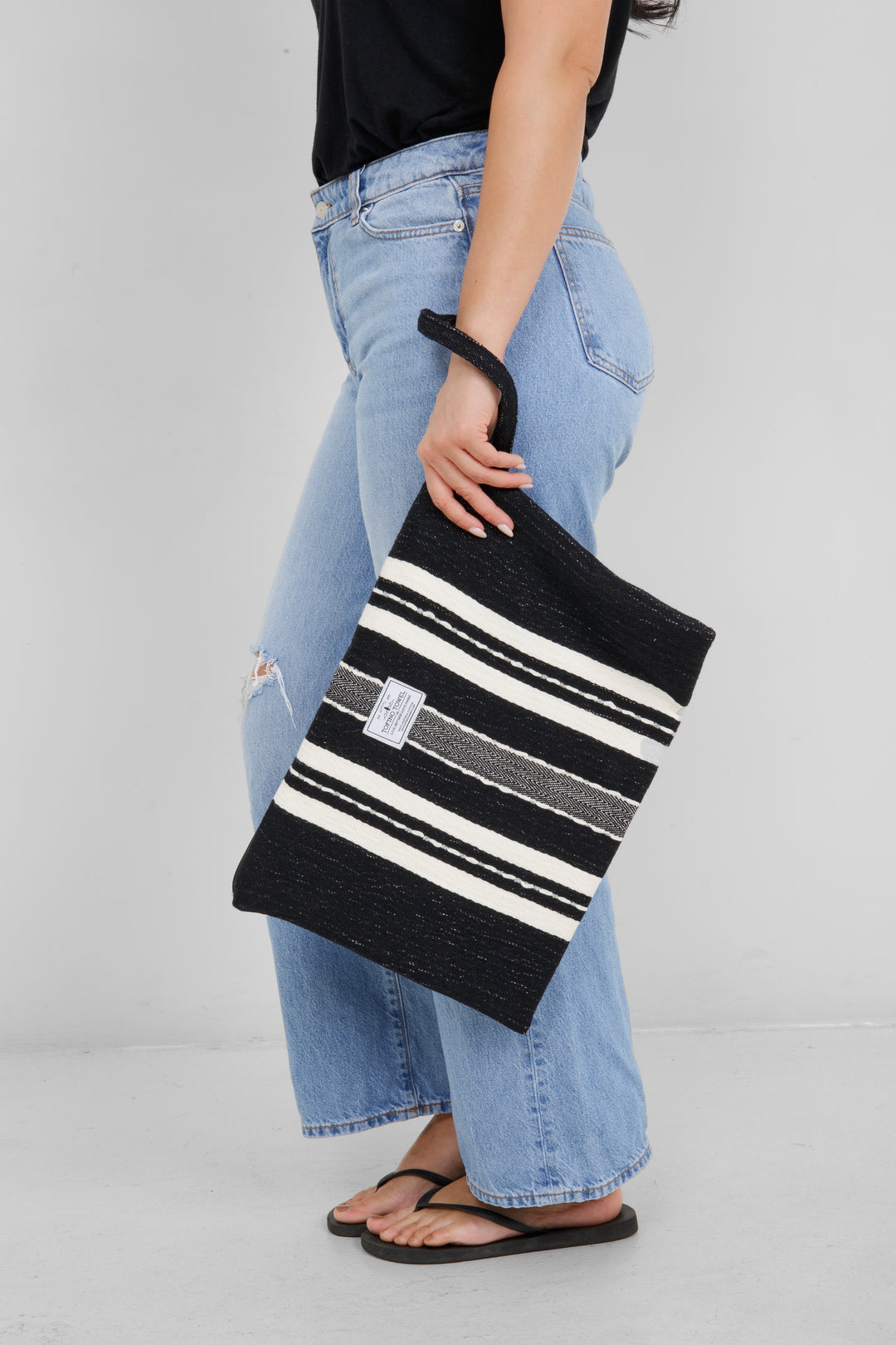 Woman holding in light blue jeans and a black t shirt holding a Large black zip up pouch with white stripes and a wrist handle on a white background
