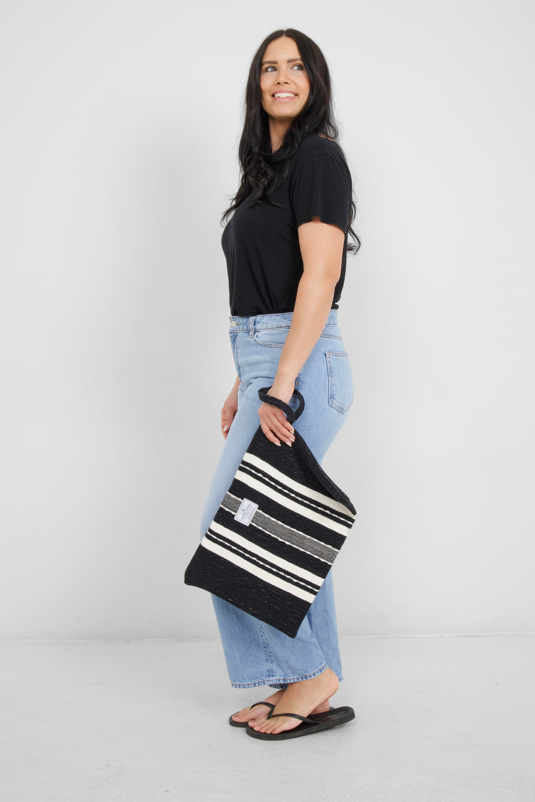 Woman holding in light blue jeans and a black t shirt holding a Large black zip up pouch with white stripes and a wrist handle on a white background