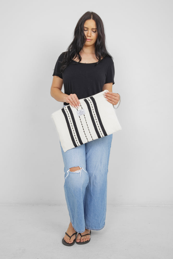 Woman holding in light blue jeans and a black t shirt holding a Large white zip up pouch with black stripes and a wrist handle on a white background