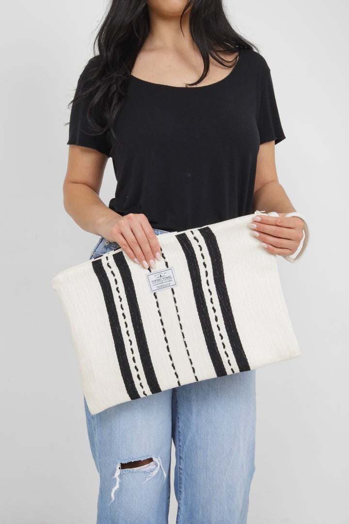 Woman holding in light blue jeans and a black t shirt holding a Large white zip up pouch with black stripes and a wrist handle on a white background