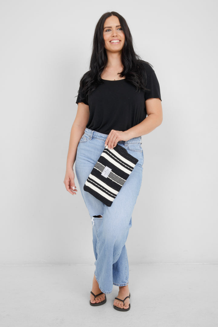 Woman wearing a black top and light blue jeans, holding a black zip up pouch with off white stripes and a wrist handle  on a grey background