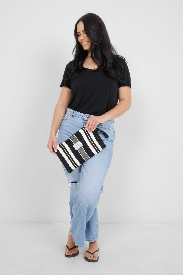 Woman wearing a black top and light blue jeans, holding a black zip up pouch with off white stripes and a wrist handle  on a grey background