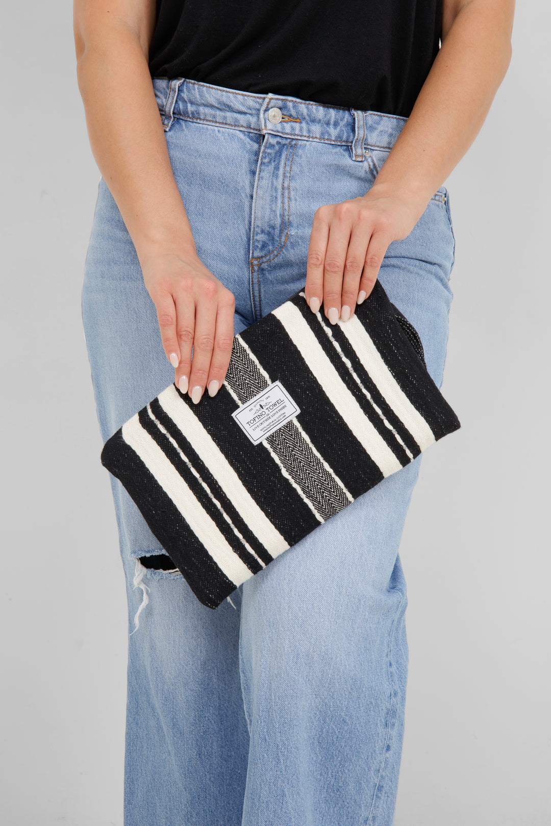 Woman wearing a black top and light blue jeans, holding a black zip up pouch with off white stripes and a wrist handle  on a grey background