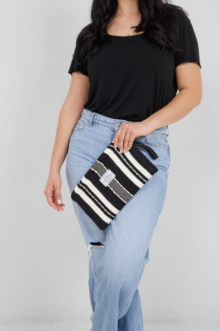 Woman wearing a black top and light blue jeans, holding a black zip up pouch with off white stripes and a wrist handle  on a grey background