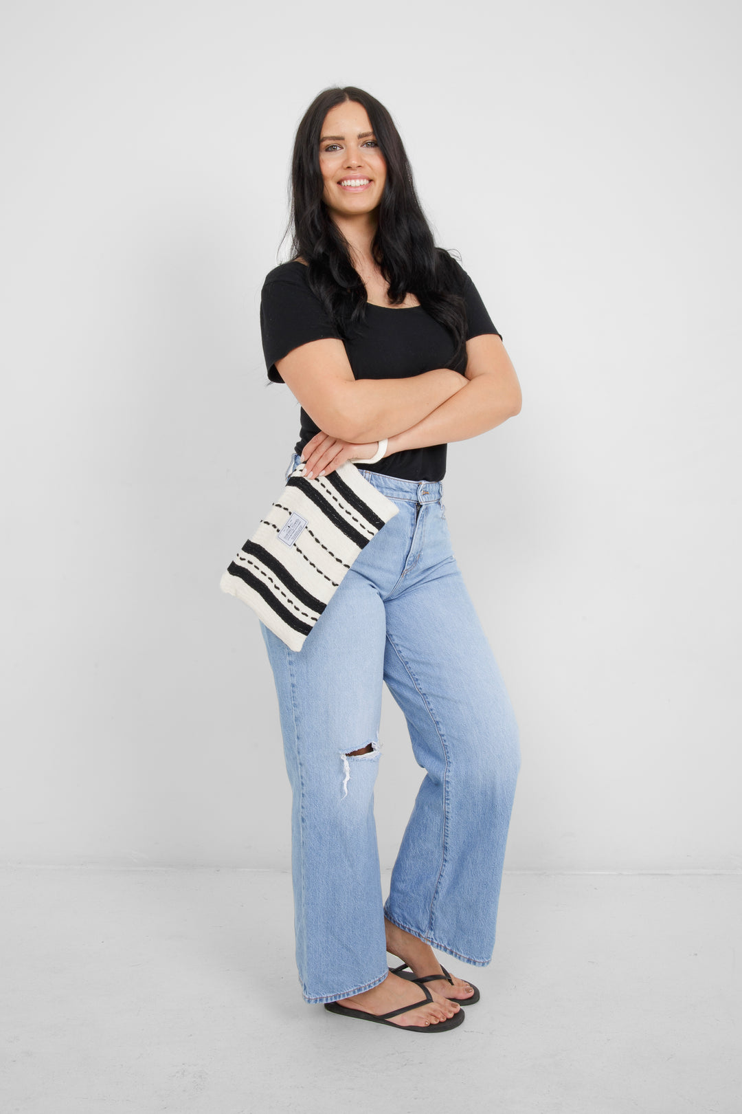 Woman wearing a black top and light blue jeans, holding a White zip up pouch with black stripes and a wrist handle  on a grey background