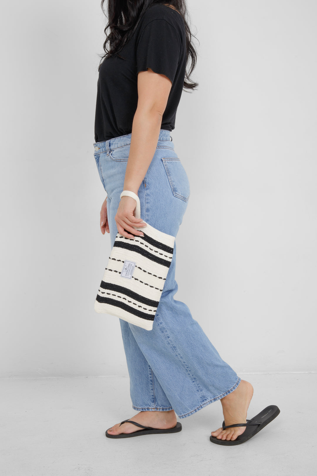 Woman wearing a black top and light blue jeans, holding a white zip up pouch with black stripes and a wrist handle  on a grey background
