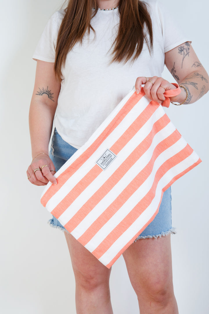 woman in white t-shirt and jean shorts holding a large white and coral striped zip up pouch with wrist handle on a white background #colour_coral-stripes