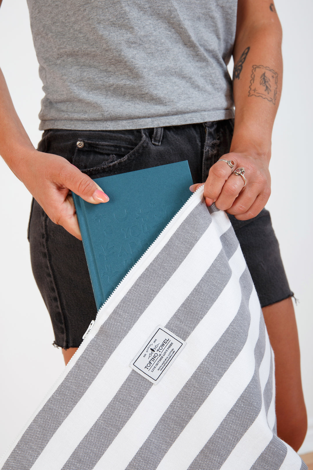 woman in grey t-shirt and black denim shorts putting a book into a large white and grey striped zip up pouch with wrist handle on a white background #colour_grey-stripes