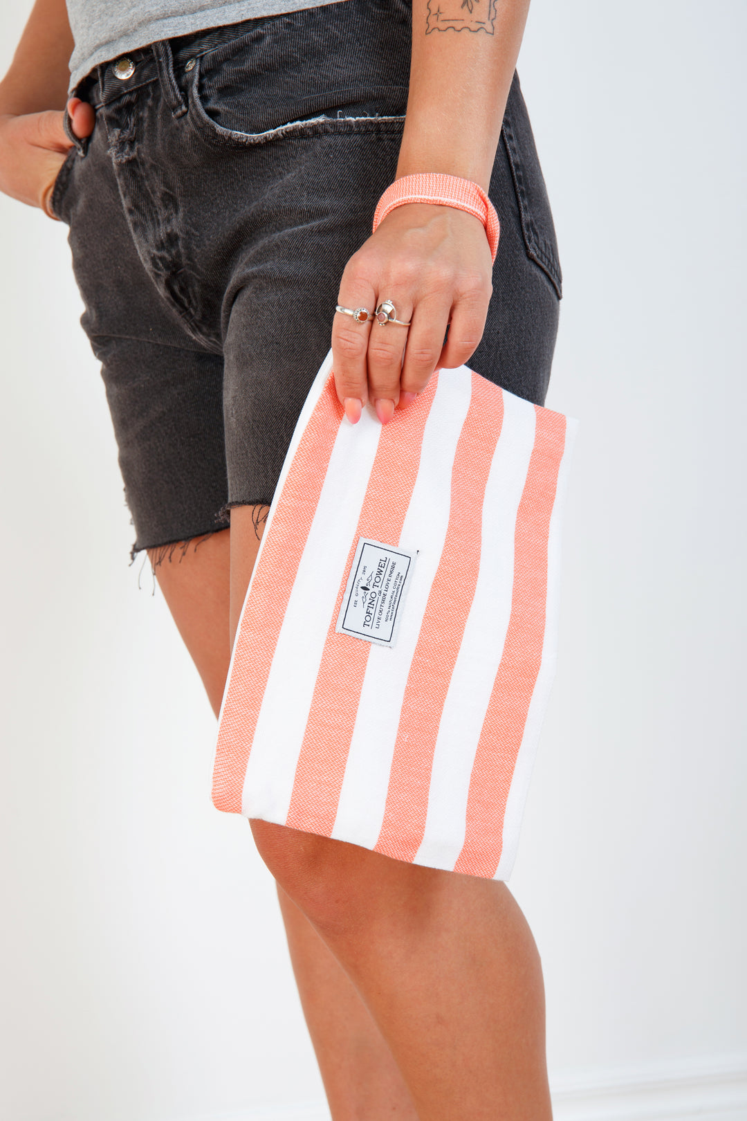 woman in grey t-shirt and black denim shorts holding a white and coral striped zip up pouch with wrist handle on a white background #colour_coral-stripes