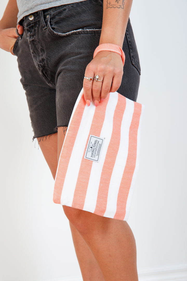 woman in grey t-shirt and black denim shorts holding a white and coral striped zip up pouch with wrist handle on a white background #colour_coral-stripes