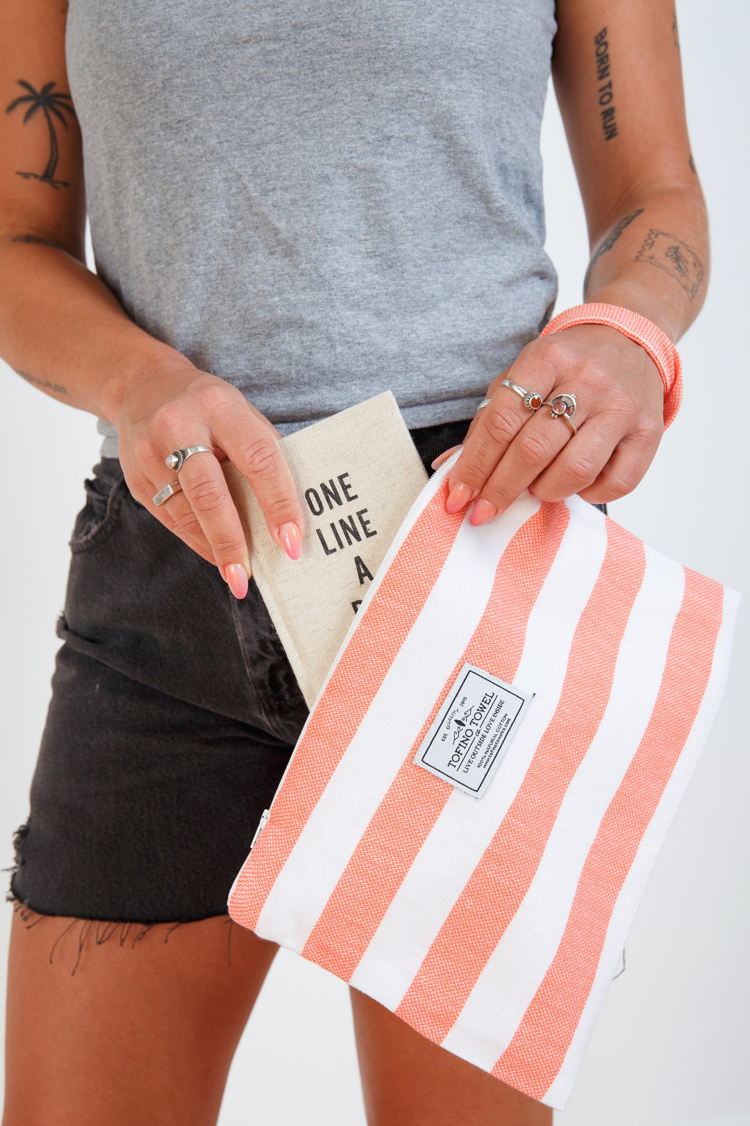 woman in grey t-shirt and black denim shorts putting a book into a white and coral striped zip up pouch with wrist handle on a white background #colour_coral-stripes