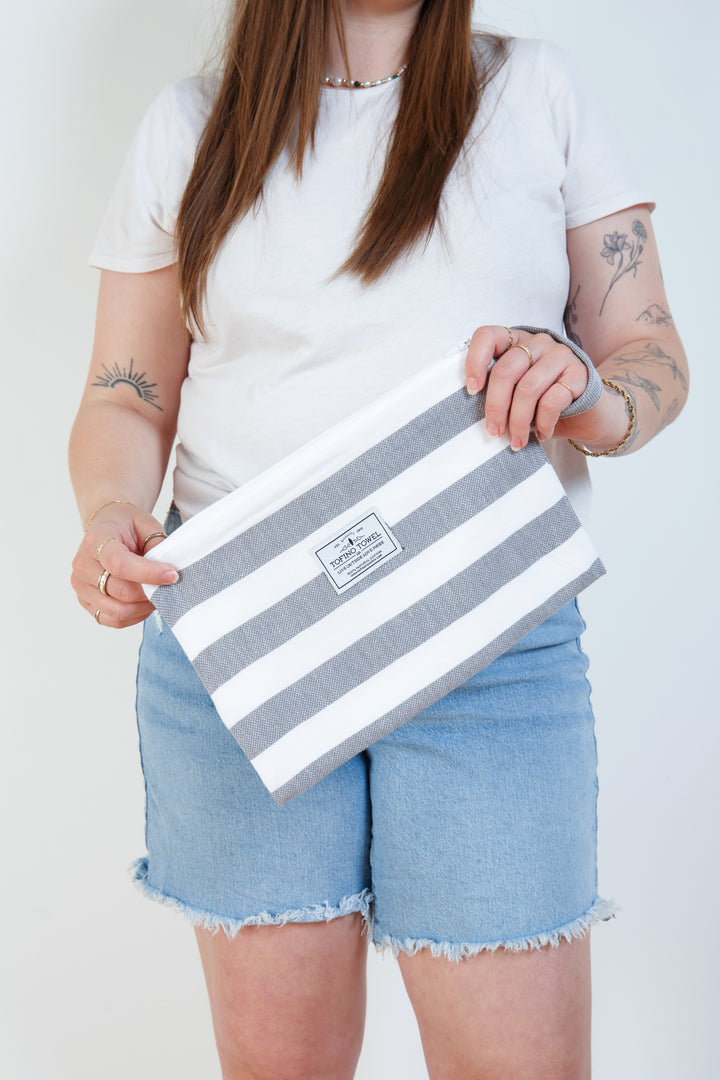 woman in jean shorts and a white t shirt holding small white and grey striped zip up pouch with wrist handle on a white background #colour_grey-stripes