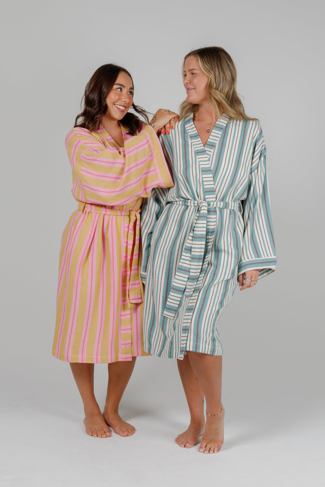 two women wearing robes, and holding a matching towel, one woman wearing a light orange robe with light and dark pink stripes and the other woman wearing an off white robe with blue and grey stripes, both standing in front of a plain light grey background #colour_ocean-mix