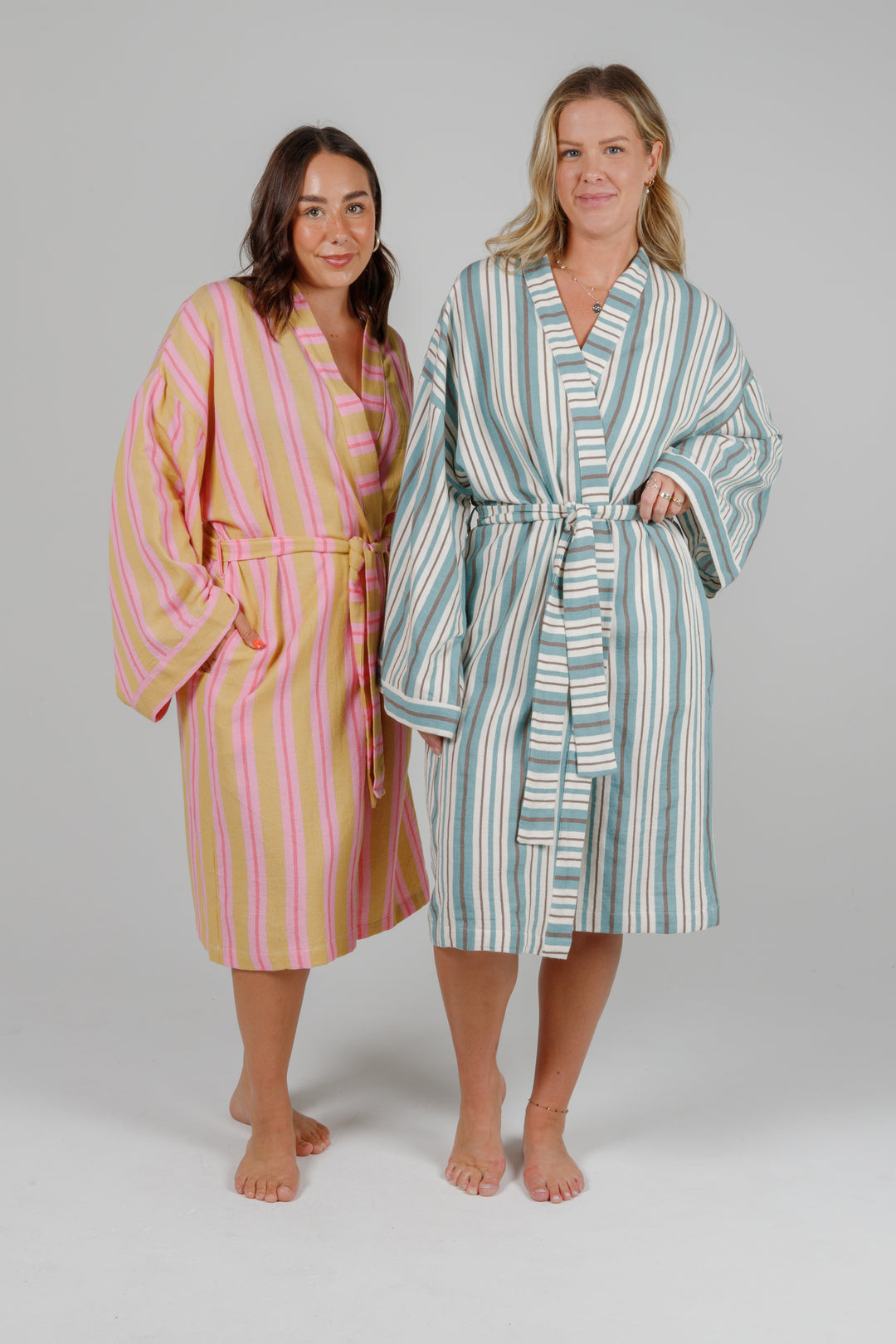 two women wearing robes, one woman wearing a light orange robe with light and dark pink stripes and the other woman wearing an off white robe with blue and grey stripes, both standing in front of a plain light grey background #colour_ocean-mix