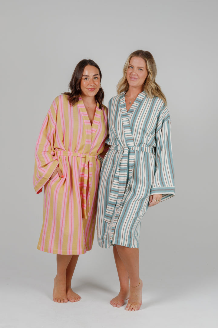 two women wearing robes, one woman wearing a light orange robe with light and dark pink stripes and the other woman wearing an off white robe with blue and grey stripes, both standing in front of a plain light grey background 
 #colour_sunset-mix