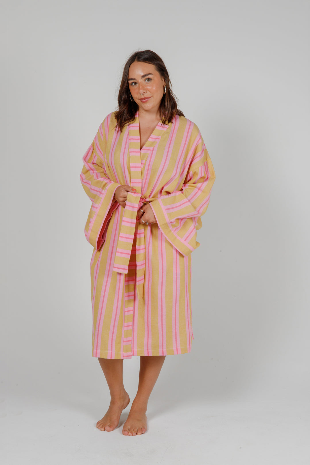 women wearing a light orange robe with light and dark pink stripes standing in front of a plain light grey background #colour_sunset-mix