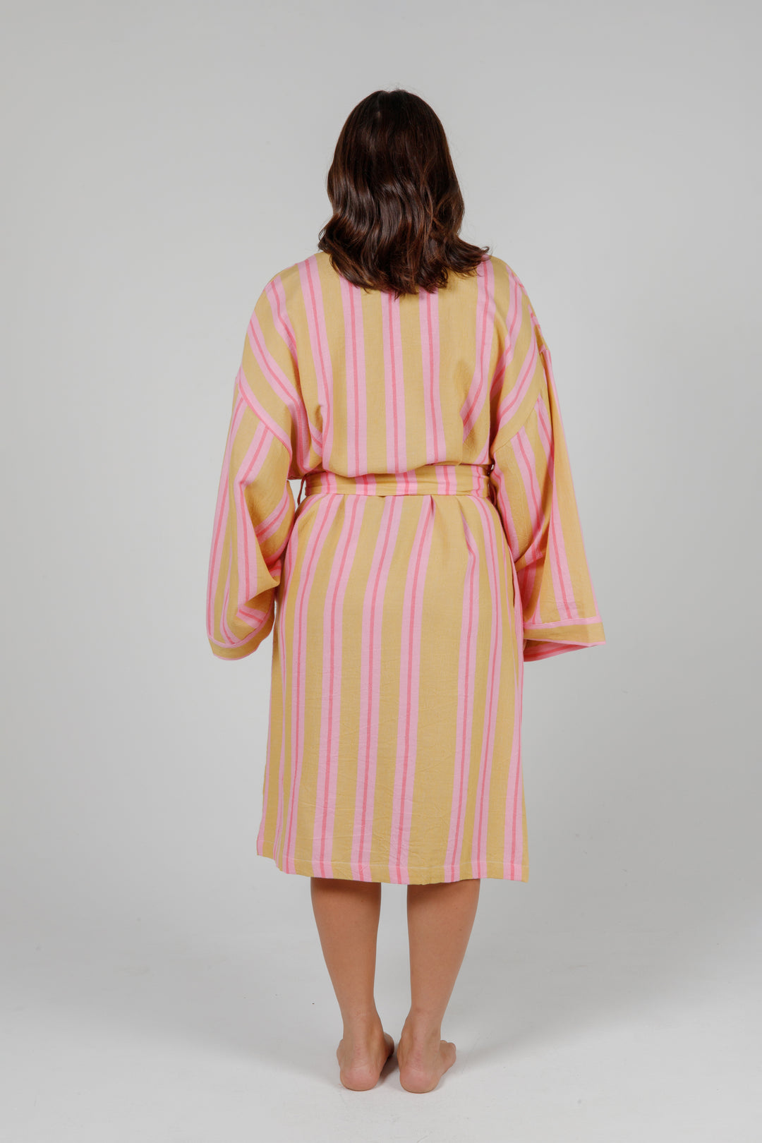 women wearing a light orange robe with light and dark pink stripes standing in front of a plain light grey background #colour_sunset-mix