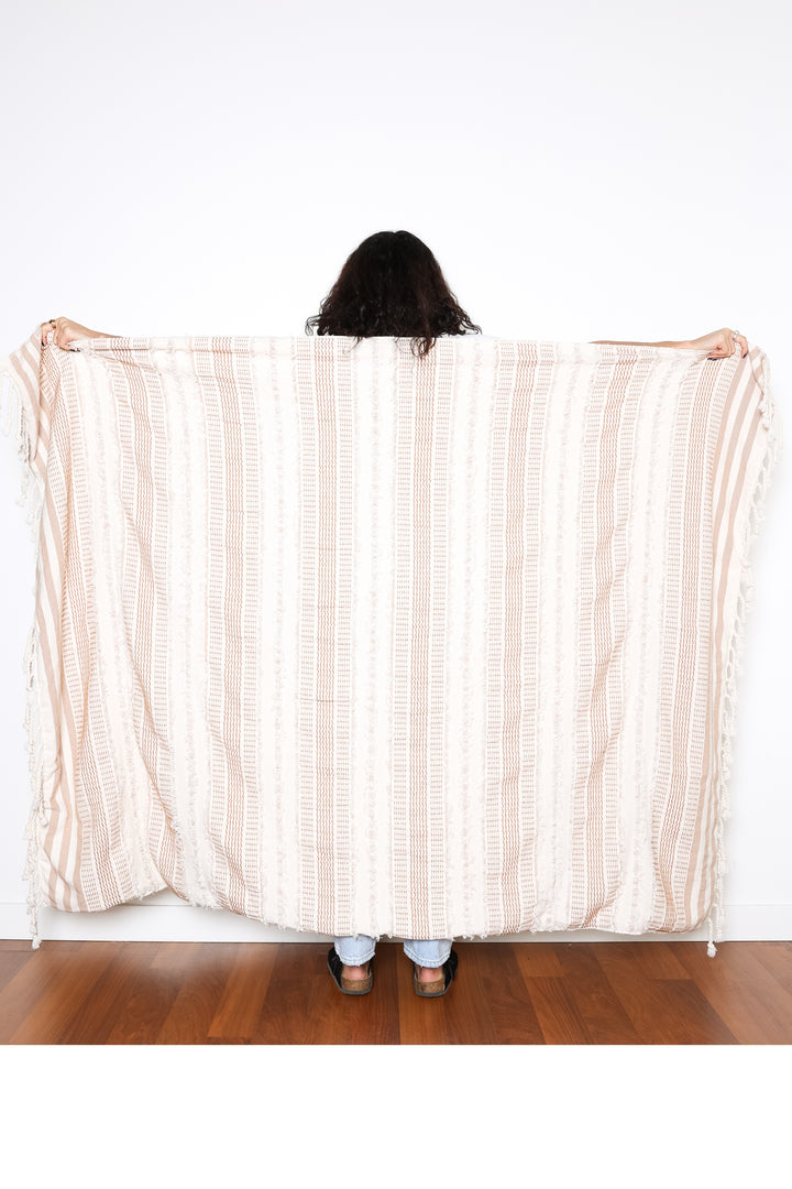 women standing backwards holding out a cream and light brown fleece lined throw with textured stripes and tassels behind her #colour_camel-stripe