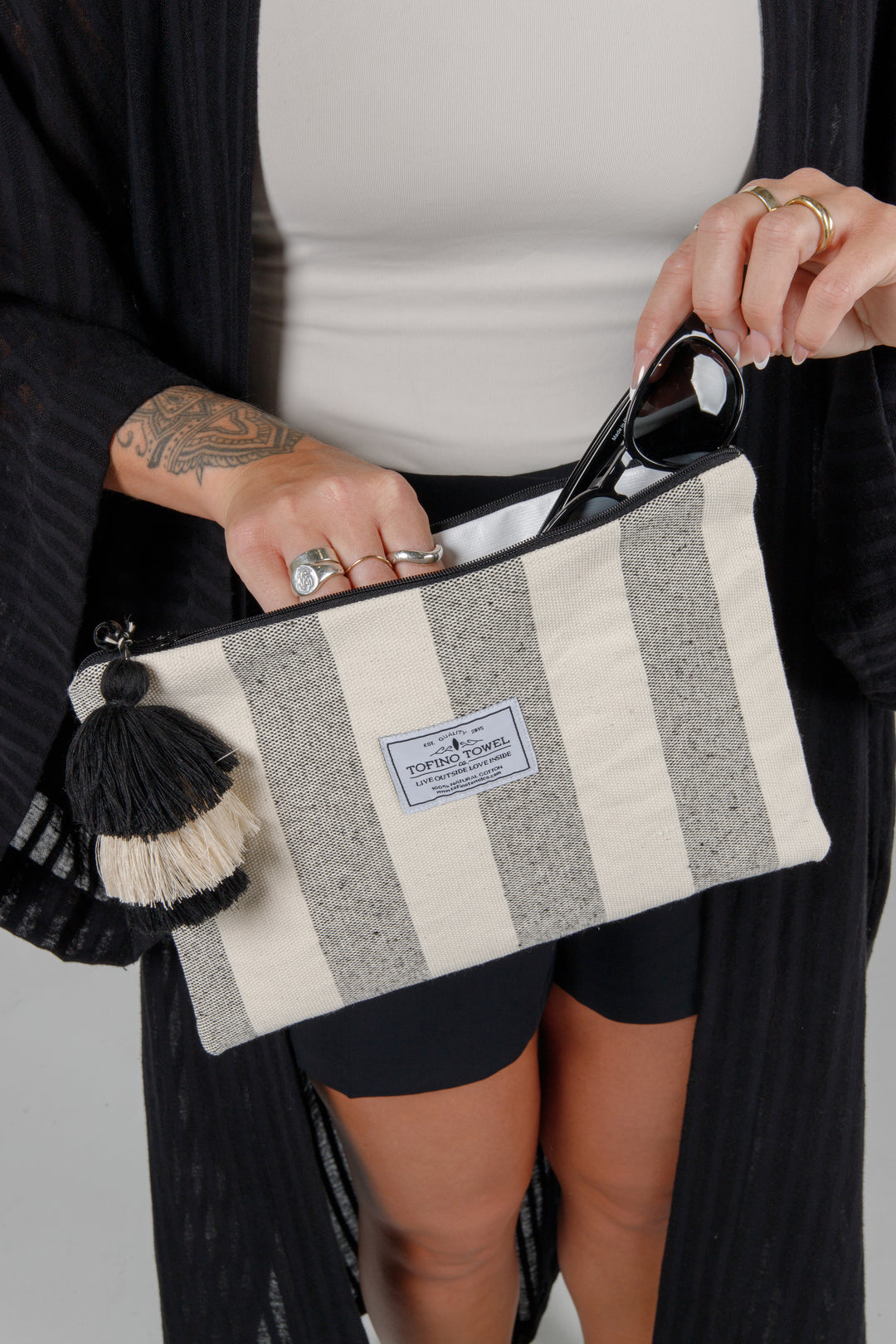 Person holding a speckled grey and off white striped canvas toiletry bag / pouch / clutch  with a tasseled zipper and sunglasses against a plain background