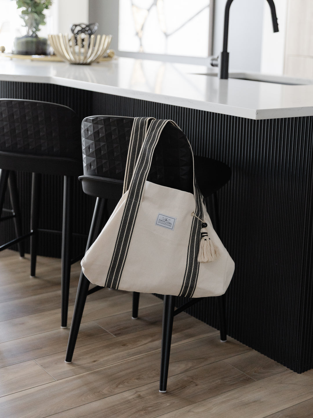 Large off white canvas tote bag with black striped straps and a large tassel hanging off the back of a black counter stool in a modern kitchen