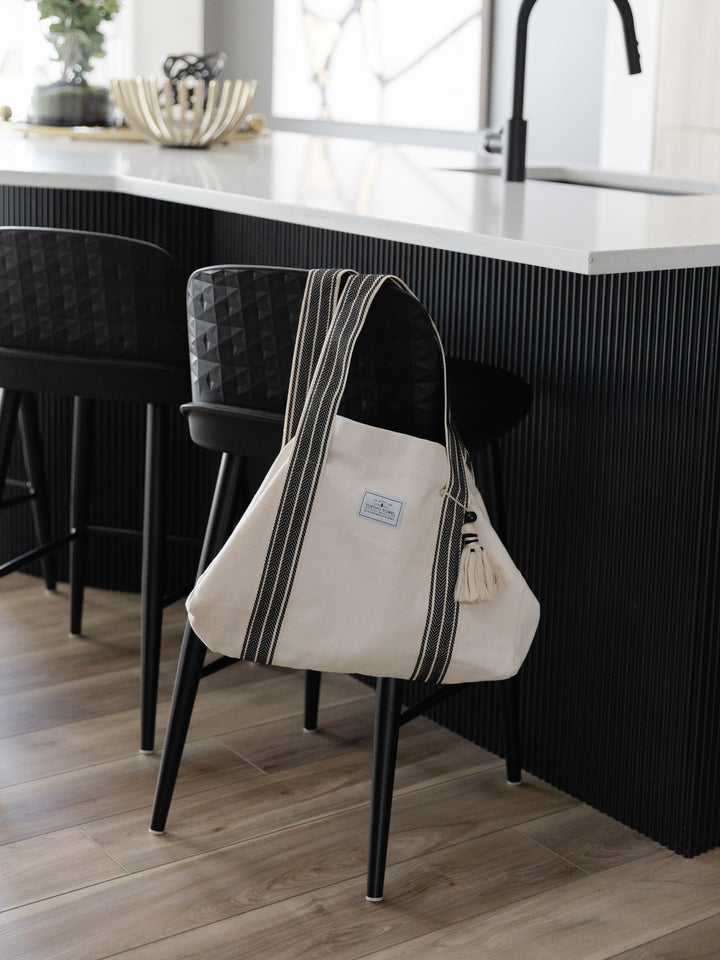 Large off white canvas tote bag with black striped straps and a large tassel hanging off the back of a black counter stool in a modern kitchen