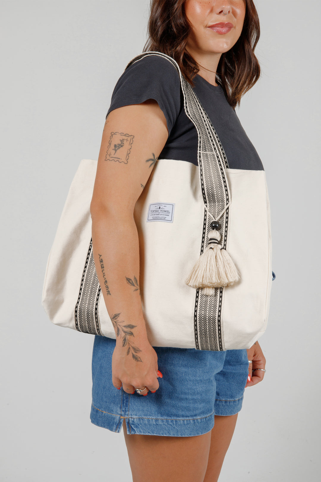 model wearing jean shorts and a black t-shirt holding a Large off white canvas tote bag with black striped straps and a large tassel on a grey background