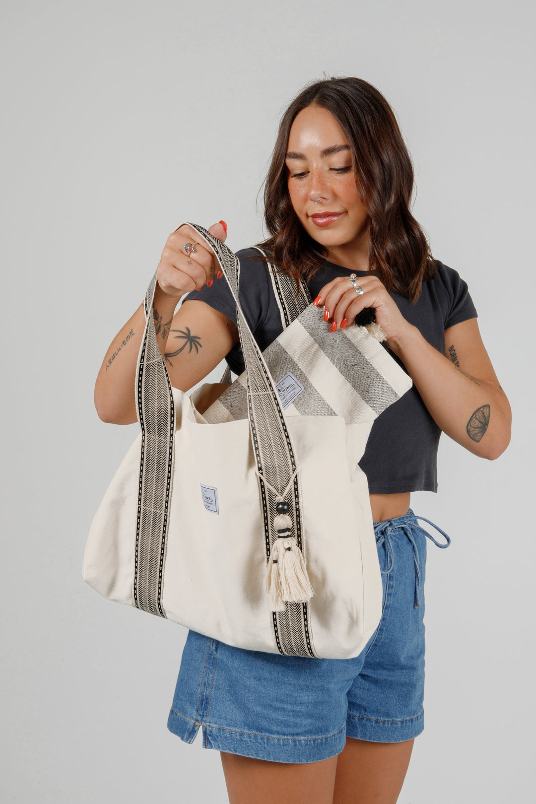 model wearing jean shorts and a black t-shirt putting a grey and off white striped pouch into  a Large off white canvas tote bag with black striped straps and a large tassel on a grey background