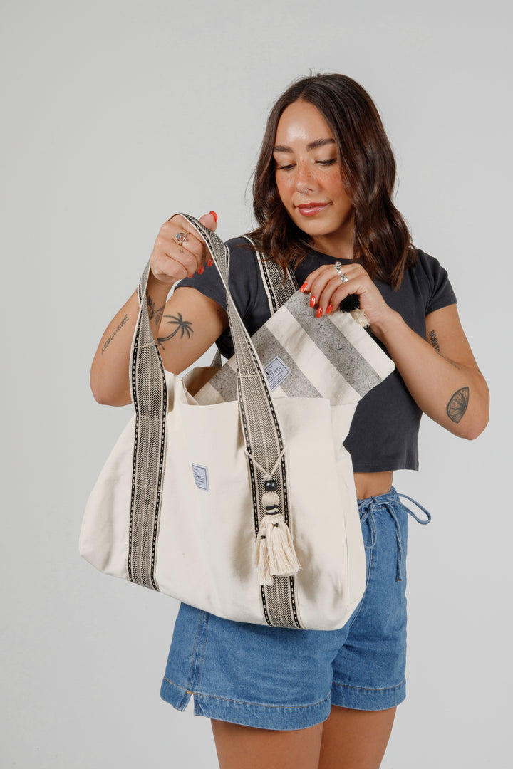 model wearing jean shorts and a black t-shirt putting a grey and off white striped pouch into  a Large off white canvas tote bag with black striped straps and a large tassel on a grey background