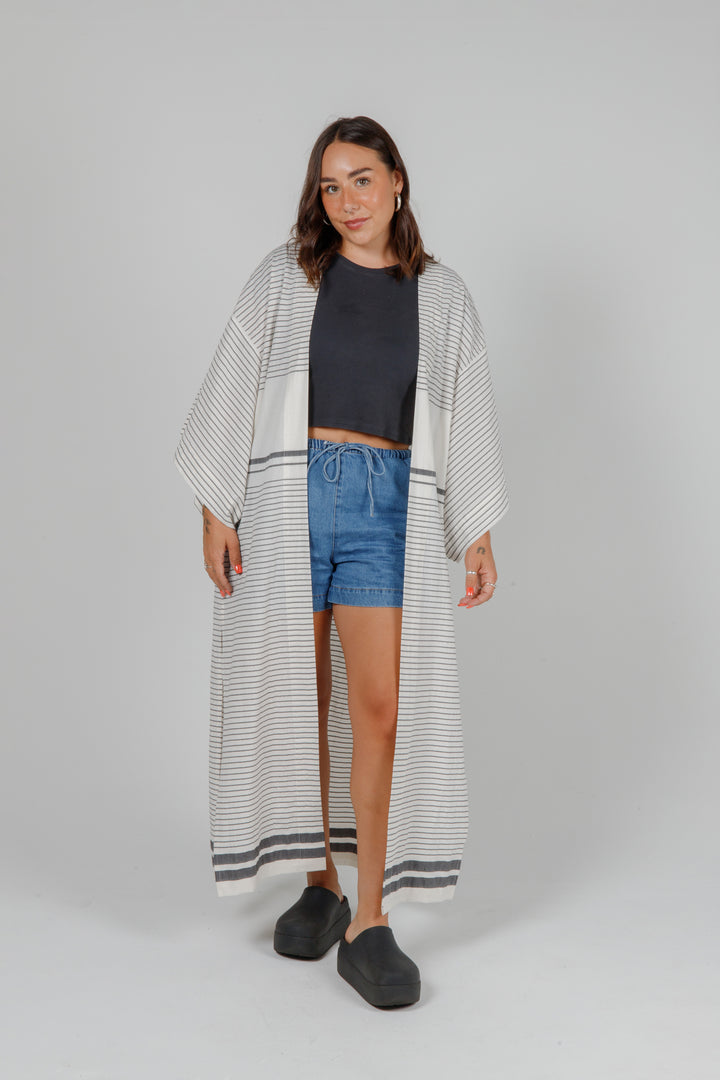 Woman wearing a long, lightweight off white striped open cover-up/robe with grey stripes and side slits over casual clothing on a plain background